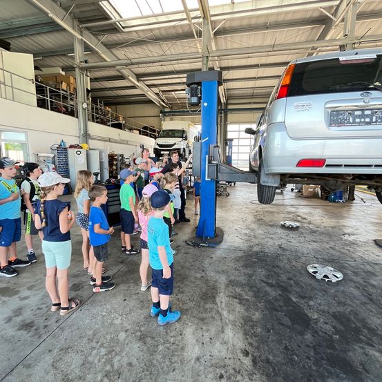 Eine Gruppe von Kindern beobachtet einen Mechaniker, der ein Auto mit einem Wagenheber in einer Garage anhebt.