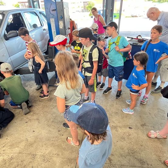 Eine Gruppe von Kindern steht in einer Garage und beobachtet ein Auto auf einem Heber. Sie tragen Freizeitkleidung und einige tragen Mützen. Eine Frau und ein älterer Mann stehen in der Nähe des Autos.