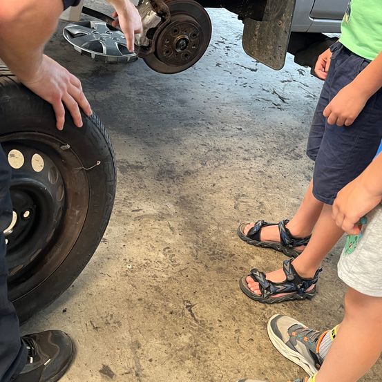 Eine Person untersucht einen Autoreifen und hält ein Teil, während zwei Kinder in Sandalen beobachten. Das Autorad ist abgenommen.