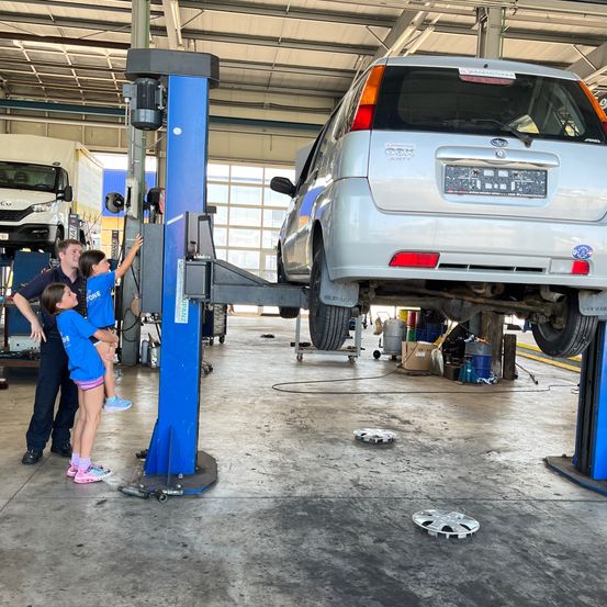 Eine Familie ist in einer Autowerkstatt. Zwei Kinder heben ein Auto an, während ein Mann hilft. Das Auto wird von einer blauen Maschine angehoben.