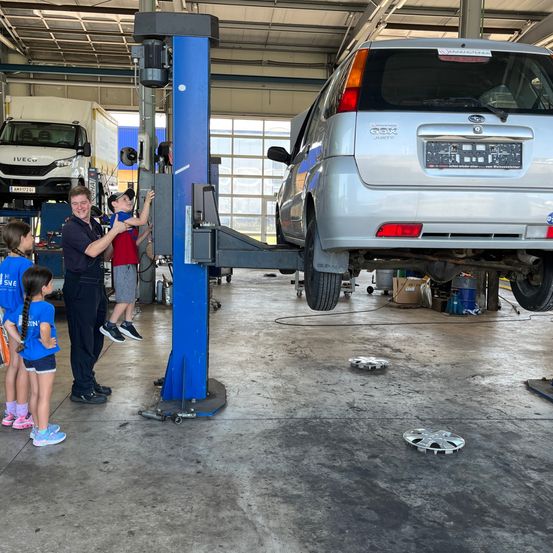Ein Mann und vier Kinder befinden sich in einer Garage, in der ein Auto angehoben wird. Das Auto ist ein silberner SUV mit einem Nummernschild, das GX8 liest.