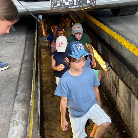 Eine Gruppe von Kindern, darunter ein Junge in einem blauen T-Shirt, geht durch einen Tunnel unter einem Auto mit dem Kennzeichen 'FORDTRUCKS'.