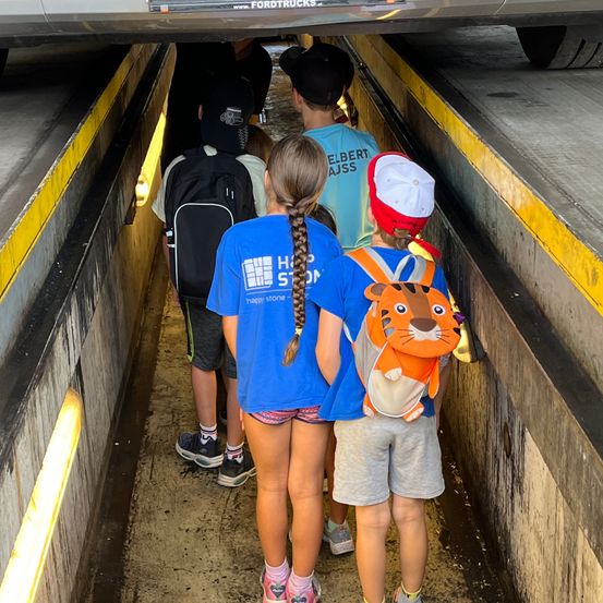 Kinder gehen durch einen Tunnel unter einem Fahrzeug mit Ford Trucks Logo. Sie tragen Rucksäcke, darunter einer mit Tigerdesign. Der Tunnel ist mit gelben und weißen Markierungen versehen.