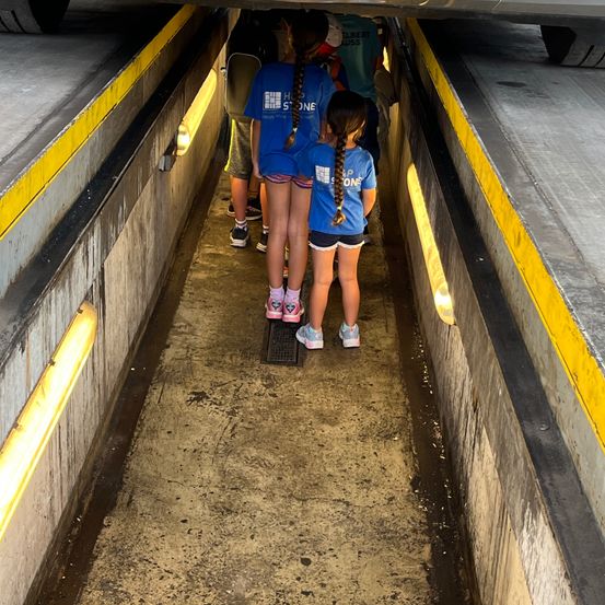 Eine Gruppe von Kindern in blauen T-Shirts geht durch einen Tunnel mit gelben Linien und Betonwänden. Ein Auto fährt über ihnen.