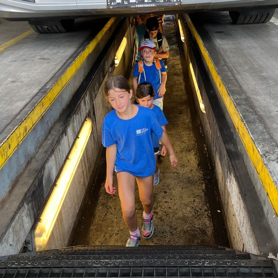 Mehrere Kinder gehen unter einem Auto in einem Tunnel. Ein Mädchen vorn trägt ein blaues T-Shirt und Turnschuhe. Dahinter folgen ein Junge mit Hut und ein weiteres Kind.