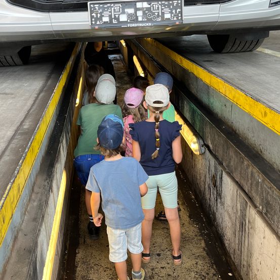 Eine Gruppe von Kindern mit Mützen geht durch einen Tunnel unter einem Fahrzeug. Der Tunnel ist eng, und die Wände sind gelb und grau. Oben der Stoßfänger eines Autos mit einem Nummernschild, das Ford Trucks liest.