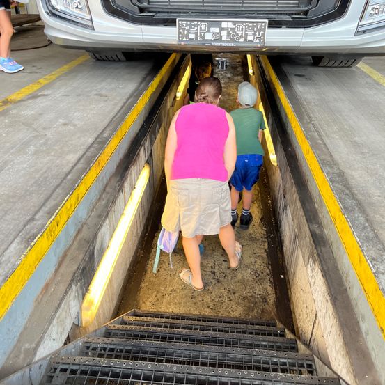 Eine Frau und zwei Kinder klettern eine Treppe unter einem Fahrzeuglift hinunter. Die Frau trägt ein rosa Hemd und Shorts, während die Kinder grüne und blaue Kleidung tragen.