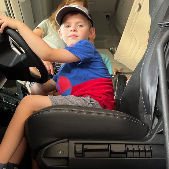 Ein junger Junge sitzt auf dem Fahrersitz eines Fahrzeugs und hält das Lenkrad. Er trägt ein blaues und rotes Shirt. Hinter ihm beobachtet ein Erwachsener.