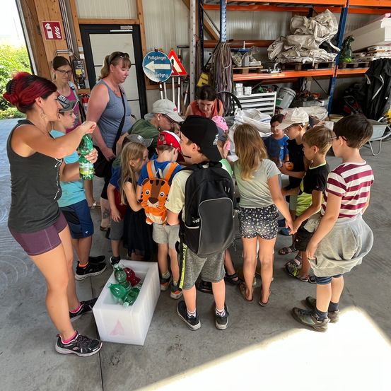 Eine Gruppe von Kindern und Erwachsenen versammelt sich in einem Raum. Eine Frau hält eine Flasche und einen Behälter. Einige Kinder haben Rucksäcke und andere halten Flaschen.