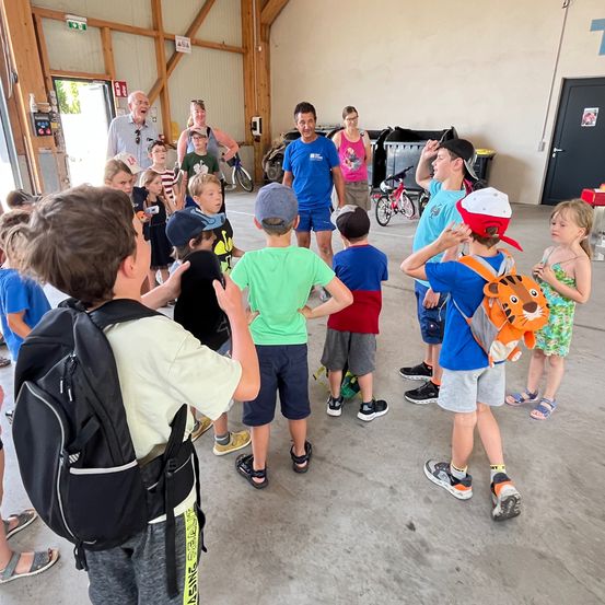 In einer Garage stehen eine Gruppe von Kindern und Erwachsenen im Kreis. Die Erwachsenen lächeln und einige halten Kameras. Ein Fahrrad ist in der Nähe geparkt. Die Kinder tragen Mützen und Turnschuhe.