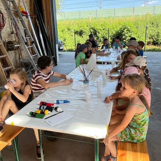 Kinder sitzen um einen langen Tisch herum und sind in einer Malaktivität beschäftigt. Sie benutzen Pinsel und Becher mit Farbe. Ein Mädchen trinkt aus einem Becher, während ein anderes Mädchen zusieht.