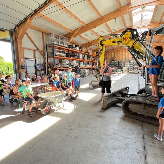 In einer Werkstatt sitzen eine Gruppe von Kindern und lauschen aufmerksam einer Frau, die in der Nähe eines gelben Baggers steht. Im Hintergrund sind Regale mit verschiedenen Gegenständen zu sehen.