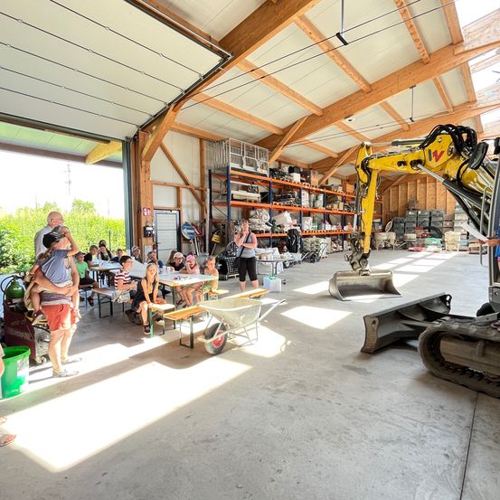 In einer großen, offenen Werkstatt sitzen mehrere Personen an Tischen, wahrscheinlich in einem Projekt engagiert. Eine Frau steht in der Nähe eines gelben Baggers. Der Raum hat Holzbalken, eine hohe Decke und Sonnenlicht strömt herein.