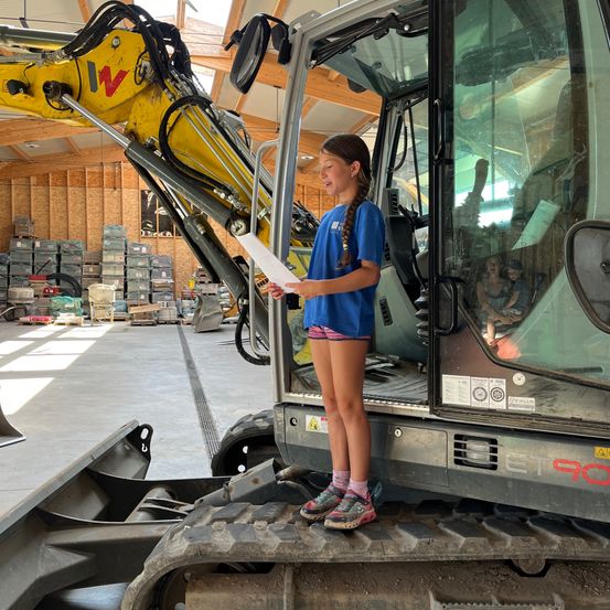 Ein junges Mädchen liest ein Papier, während sie auf einem großen gelben Bagger in einer Lagerhalle steht. Sie trägt ein blaues Shirt und Shorts.