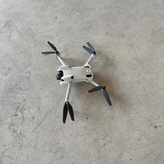 Ein kleiner weißer Drohne mit vier Propellern steht auf einer Betonfläche. Die Drohne hat ein schlankes Design mit orangefarbenen Spitzen an den Propellern.