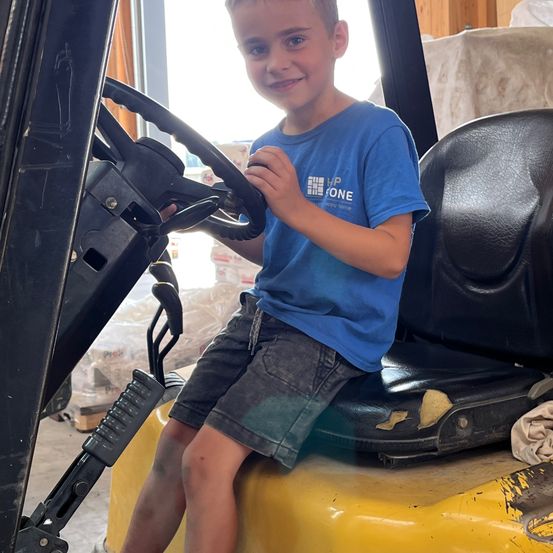 Ein junger Junge sitzt mit einem Lächeln im Gesicht in einem Gabelstapler. Er trägt ein blaues T-Shirt und graue Shorts.