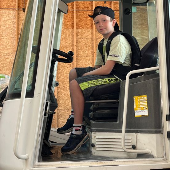 Ein Junge mit einem Rucksack sitzt auf dem Fahrersitz eines weißen Fahrzeugs, möglicherweise eines Baufahrzeugs. Das Fahrzeug ist offen, und die Füße des Jungen sind auf dem Boden.