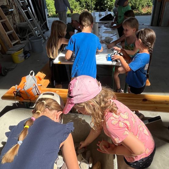 Eine Gruppe von Kindern und Erwachsenen arbeitet gemeinsam an einem Projekt in einer Werkstatt. Die Kinder tragen Mützen und einige sitzen auf Stühlen.