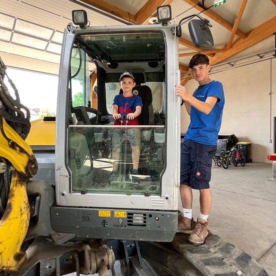 Zwei junge Jungen stehen neben einem großen Fahrzeug. Ein Junge sitzt im Fahrzeug, während der andere auf dem Reifen des Fahrzeugs steht. Der Junge im Fahrzeug hält das Lenkrad.