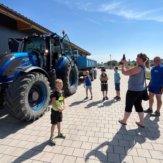 Eine Frau macht ein Foto von einer Gruppe von Kindern in der Nähe eines Traktors und eines Lastwagen im Freien.