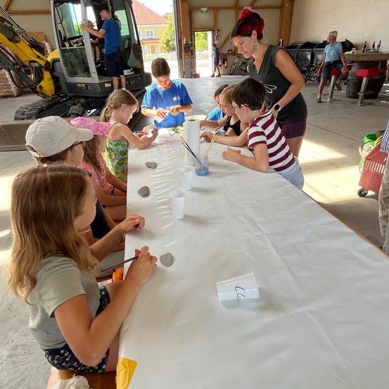 Eine Gruppe von Kindern sitzt um einen Tisch herum und bemalt Steine mit Bechern und Pinseln. Eine Frau in einem ärmellosen Oberteil beaufsichtigt sie, während ein Mann in einem gelben Baufahrzeug sitzt.