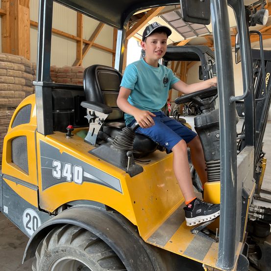 Ein junger Junge sitzt in einem gelben Gabelstapler mit der Aufschrift 340, trägt einen Hut, ein grünes T-Shirt und blaue Shorts und hat ein lächelndes Gesicht.