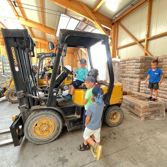 Drei junge Jungen spielen mit einem Gabelstapler in einem Lagerhaus. Ein Junge sitzt im Gabelstapler, während andere stehen und ihn anschauen. Das Lagerhaus ist mit gestapelten Paletten gefüllt.