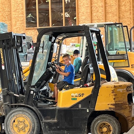 Zwei Jungen sitzen in einem gelben Gabelstapler, einer fährt. Dahinter eine Holzwand und ein großes Fenster.