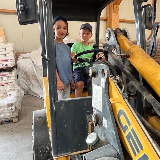 Zwei junge Jungen sitzen in einem gelben Traktor in einem Lagerhaus und lächeln. Ein Stapel Profi-Markensäcke steht hinter ihnen.