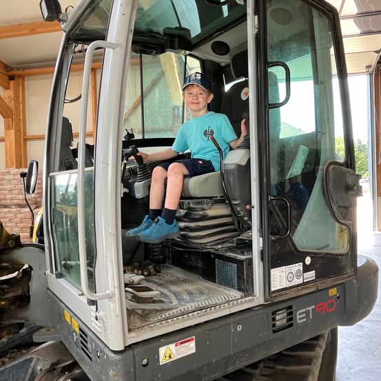 Ein junger Junge sitzt auf dem Fahrersitz eines großen, grauen Baufahrzeugs mit 'ET90' an der Seite. Er trägt ein blaues Shirt und eine Kappe.