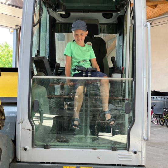Ein junger Junge sitzt in der Kabine eines Baufahrzeugs. Er trägt ein grünes T-Shirt und blaue Shorts, eine Baseballkappe auf dem Kopf.