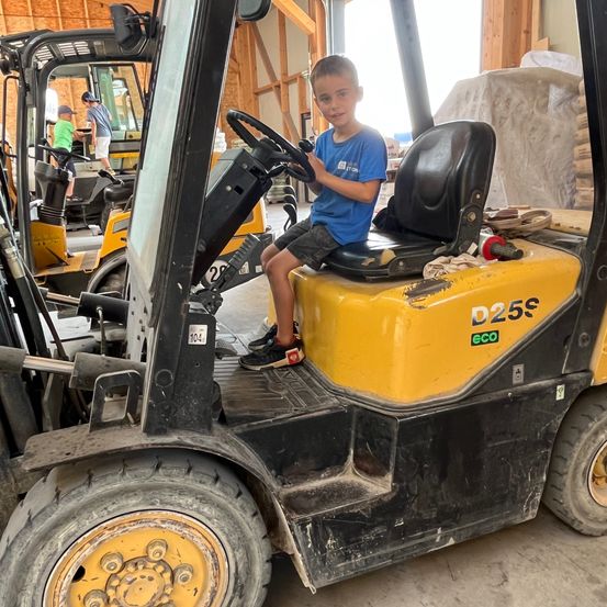 Ein junger Junge in einem blauen T-Shirt sitzt auf einem Gabelstapler in einem Lagerhaus. Hinter ihm sind zwei andere Kinder zu sehen, eines trägt einen Hut.