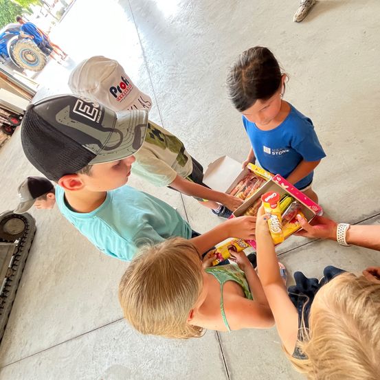 Eine Gruppe von Kindern versammelt sich um eine Box mit Snacks. Ein Kind hält eine Box, und andere greifen danach. Die Kinder tragen Freizeitkleidung. Im Hintergrund sitzt eine Person auf einem Fahrzeug.