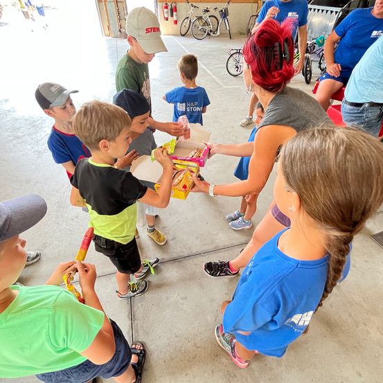 Eine Gruppe von Kindern und Erwachsenen versammelt sich in einer Garage. Eine Frau verteilt Boxen mit Snacks an die Kinder. Die Kinder halten verschiedene Gegenstände und tragen Mützen und Turnschuhe. Fahrräder und ein Feuerlöscher sind im Hintergrund zu sehen.