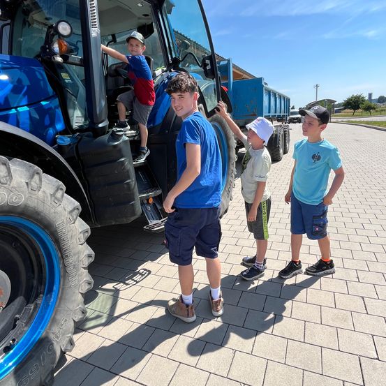 Drei Jungen stehen neben einem blauen Traktor. Einer sitzt im Inneren des Traktors, während die anderen draußen stehen. Sie tragen alle Mützen und Turnschuhe. Der Schatten des Traktors wird auf den Ziegelboden geworfen.