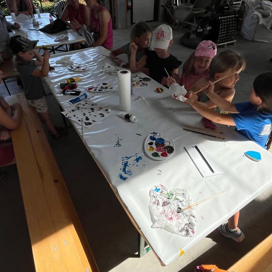 Eine Gruppe von Kindern malt auf einem weißen Tisch. Sie haben Malpinsel und Teller mit Farbe. Einige Erwachsene sitzen im Hintergrund an anderen Tischen.