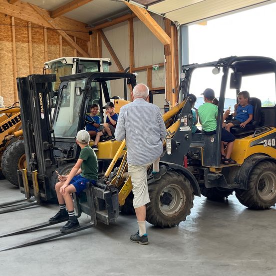 Ein älterer Mann steht vor einem Gabelstapler, auf dem Kinder sitzen. Die Kinder essen Eis. Dahinter stehen zwei weitere Gabelstapler. Der Bereich ist mit Holzwänden umschlossen.