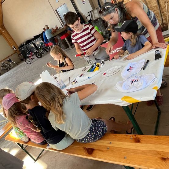 Eine Gruppe von Kindern malt auf einem Tisch mit weißer Tischdecke. Eine Frau und ein Kind schauen sich das Gemälde an. Ein Fahrrad steht in der Ecke.