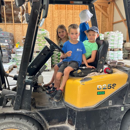 Drei Kinder sitzen in einem Gabelstapler, eines mit Hut, ein anderes isst und ein Mädchen hinter ihnen. Sie befinden sich in einem Lager mit Holzwänden und gestapelten Materialien.