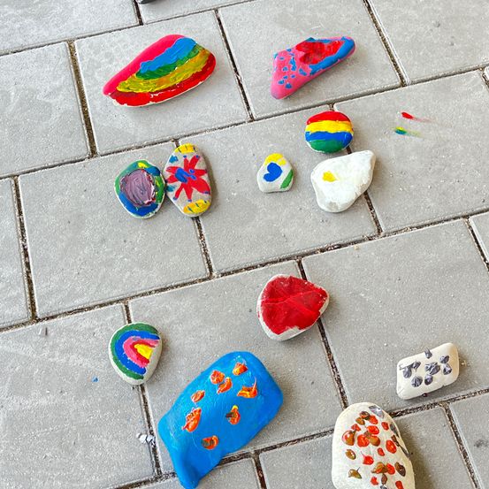 Mehrere bemalte Steine sind auf einem Ziegelpflaster angeordnet. Sie zeigen verschiedene Farben und Designs, darunter Regenbögen, Herzen und Blumen.