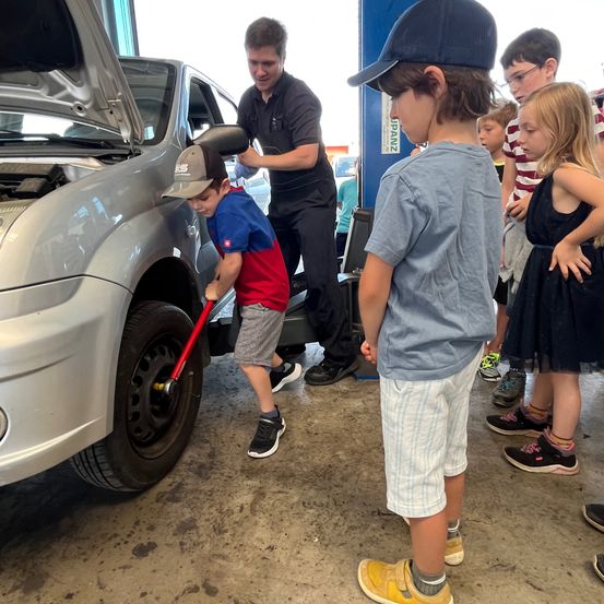 Ein Junge benutzt einen Schraubenschlüssel an einem Autoreifen, während ein Mann zuschaut. In der Nähe beobachten mehrere Kinder.