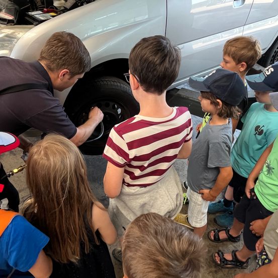 Ein Mann mit Brille überprüft ein Autoreifen, während eine Gruppe von Kindern, einige in Mützen und Sandalen, neugierig zuschauen.