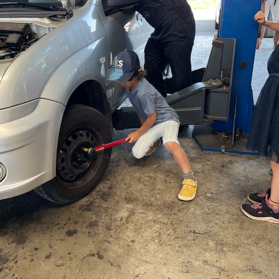 Ein Kind benutzt einen Schraubenschlüssel, um das Rad eines Autos zu festziehen, während ein Erwachsener zuschaut. Das Auto ist auf einem blauen hydraulischen Wagenheber angehoben.