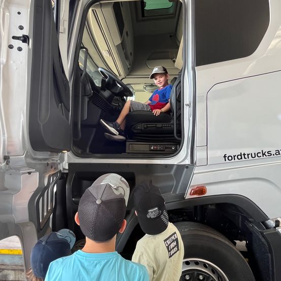 Drei Jungen erkunden einen weißen Ford-Lkw, wobei ein Junge auf dem Fahrersitz sitzt. Der Lkw hat den Text 'fordtrucks.a' an seiner Seite.