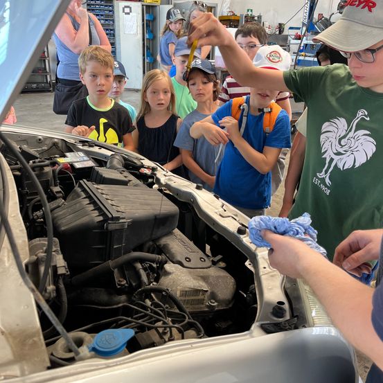 Eine Gruppe von Kindern beobachtet einen offenen Automotor, wobei ein Junge ein Werkzeug hält. Sie sind in einer Garage versammelt, und eine Frau steht in der Nähe.