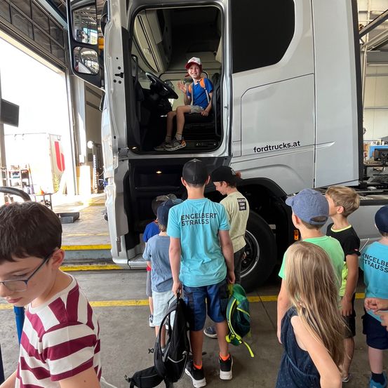 Eine Gruppe von Kindern beobachtet einen Jungen, der in einem Lastwagen sitzt, mit einer Website-Adresse an der Seite. Der Junge lächelt und winkt.