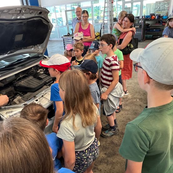 Eine Gruppe von Kindern und Erwachsenen versammelt sich um ein Auto mit geöffneter Motorhaube in einer Garage. Einige Kinder tragen Mützen, und zwei Frauen halten jeweils ein Kind. Der Motor des Autos ist sichtbar.