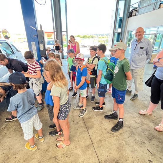 Eine Gruppe von Kindern und Erwachsenen versammelt sich in einer Garage und beobachtet ein Auto, während ein Mann mit Brille und gestreiftem Hemd in der Nähe steht.
