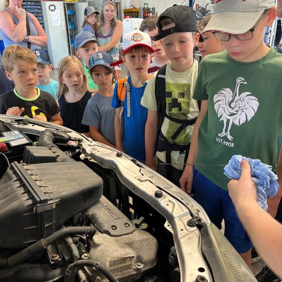 Eine Gruppe von Kindern in einer Garage untersucht einen Automotor, angeleitet von einem Erwachsenen. Die Motorhaube des Autos ist geöffnet, und der Motor ist sichtbar. Die Kinder tragen Mützen und Turnschuhe.