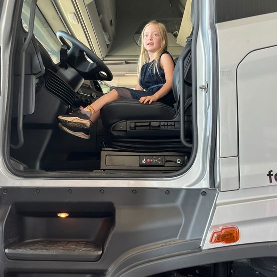 Ein junges Mädchen mit blonden Haaren sitzt auf dem Fahrersitz eines großen Fahrzeugs. Sie trägt ein Kleid und Sneakers. Das Fahrzeug hat einen silbernen Körper und eine offene Tür.